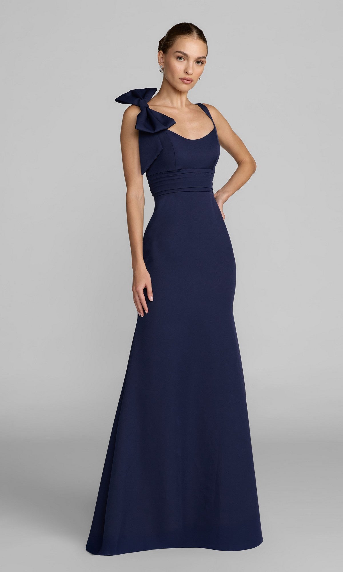 A woman stands against a plain gray background wearing the Mac Duggal 49964 long formal dress in navy—a floor-length, sleeveless gown featuring a large bow on one shoulder.